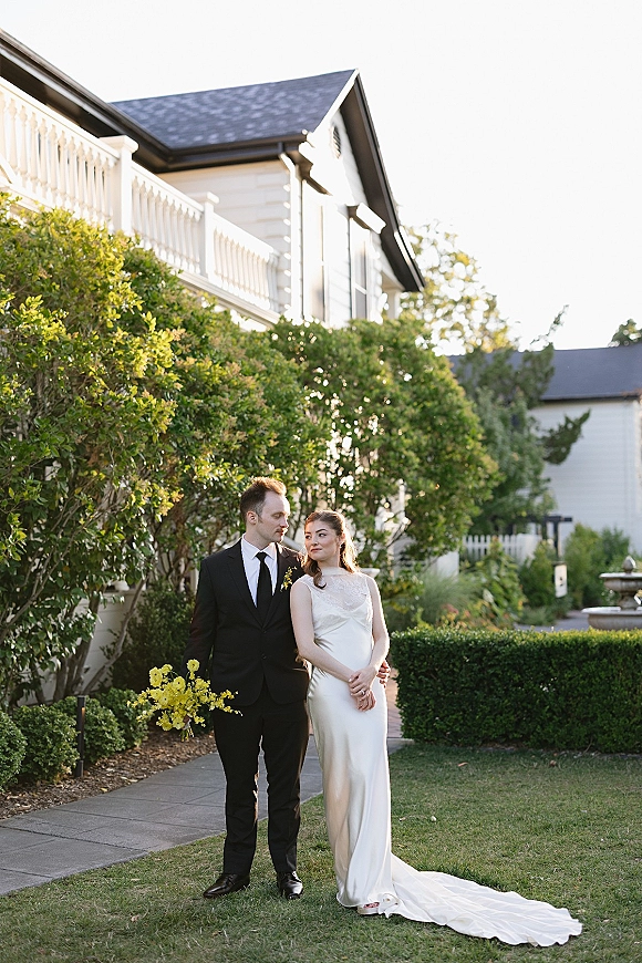 Couple portrait of bride in a minimalist wedding dress with long train and groom in black suit holding a yellow bouquet by a garden fountain