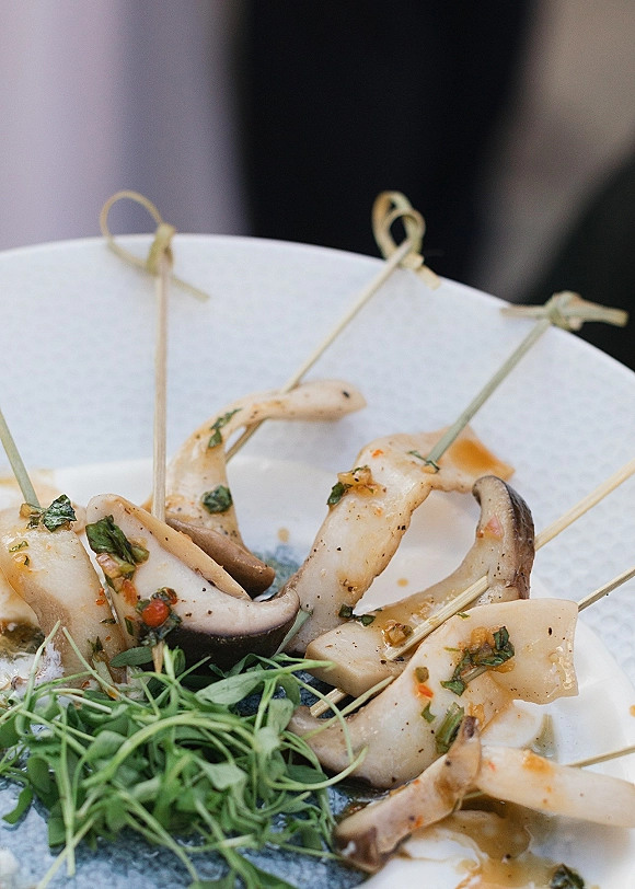 Wedding appetizers on bamboo skewers with mushrooms and microgreens, served on a ceramic plate with sauce in a blurred indoor setting