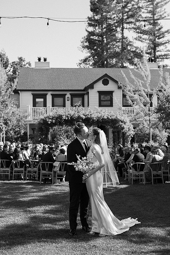 Wedding kiss portrait of bride and groom kissing, bride holding a bouquet with veil accent on an outdoor lawn amid seated guests and string lights