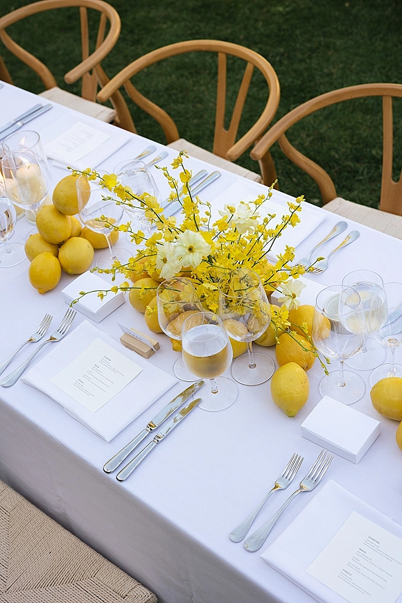 Reception tablescape with lemon wedding tablescape centerpiece of yellow flowers and lemons, set with candles, menus, wine glasses on a grass lawn