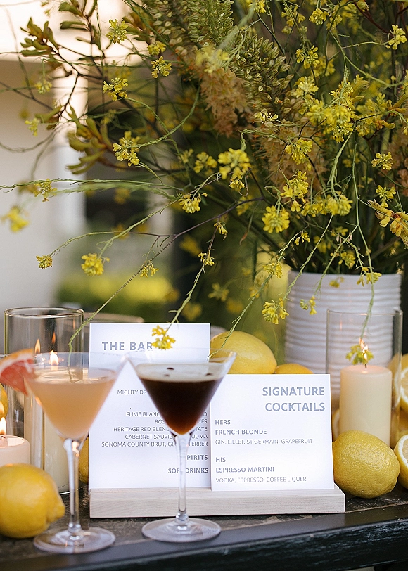 Wedding bar menu with a signature cocktails sign displayed in a holder beside coupe glasses, lemons, and candles in a blurred garden setting