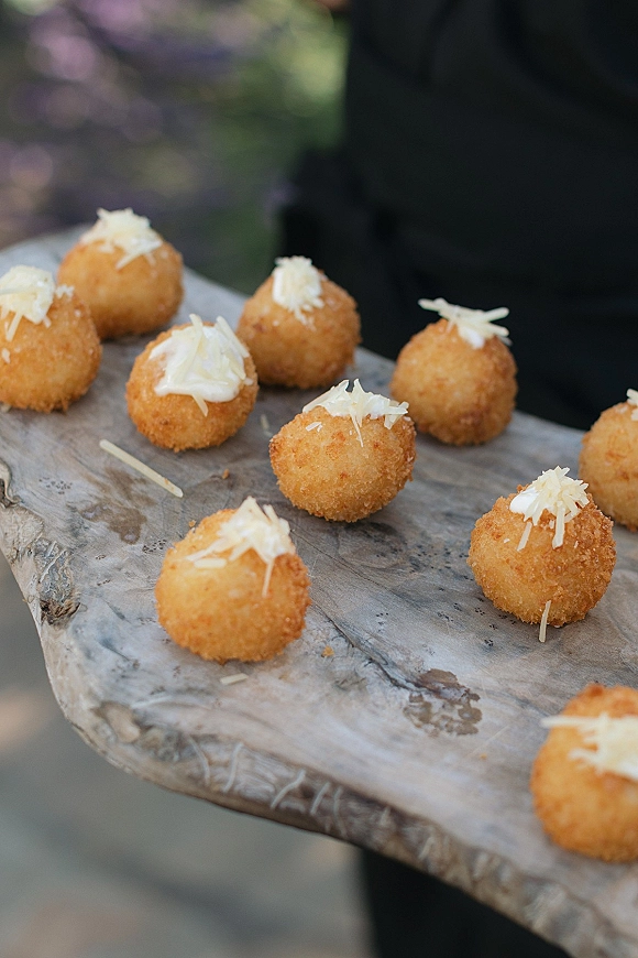 Wedding cocktail appetizers served as passed hors d'oeuvres, fried bites topped with grated cheese on a wooden board, with blurred greenery behind