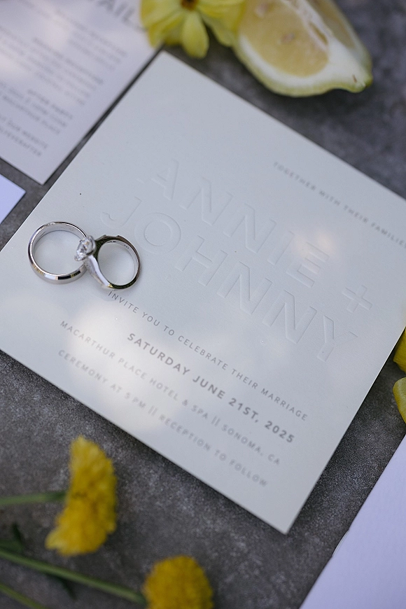 Wedding invitation suite with letterpress wedding invitation, wedding rings and lemon slice beside yellow flowers on a gray stone flat lay background