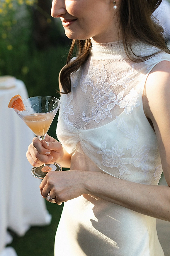 Bride portrait in a lace wedding dress lace bodice with illusion neckline, pearl studs, holding a cocktail with citrus garnish in soft greenery light