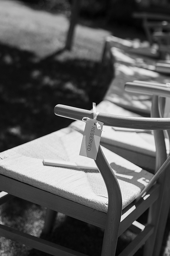 Reserved chair sign with a ribbon-tied tag on a wooden ceremony chair, set along an outdoor lawn seating row with trees and sunlit shadows