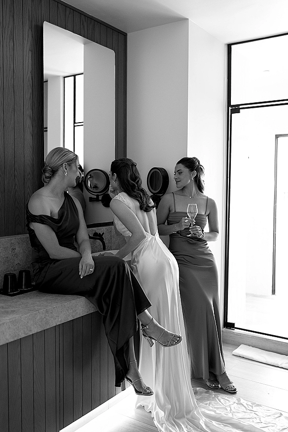Bridesmaids getting ready with a bride with bridesmaids, chatting by a bathroom vanity as one lifts a champagne flute in a toast