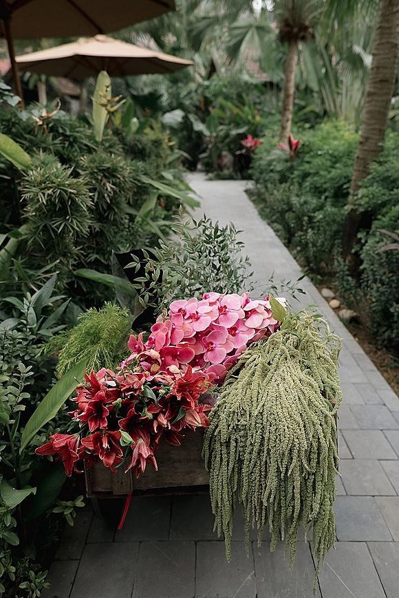 Wedding flowers wrapped in black paper with pink orchids and red lilies, held along a garden walkway with tropical palms in back