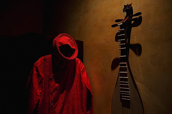 Groom sherwani in red embroidered fabric with matching turban hung on wall hooks against a beige wall with a hanging string instrument