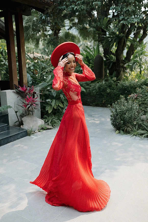 Wedding guest outfit in a red wedding guest dress with long sleeve lace and pleated skirt, paired with a wide brim hat in a tropical garden