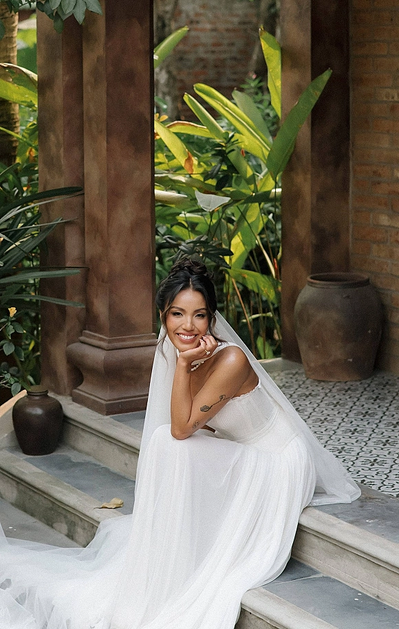Bridal portrait of a bride sitting on steps in a strapless wedding dress with veil and pearl necklace in a tropical courtyard setting