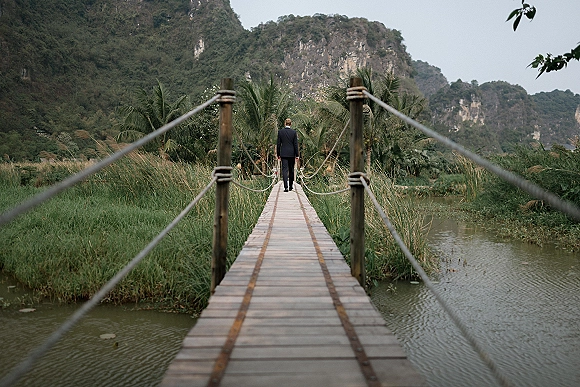 Groom portrait of a man in a black suit walking away on a wooden rope bridge over a river, with palms, mountains, and overcast sky