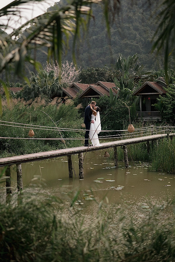 Wedding kiss portrait of bride and groom kissing on a wooden footbridge with rope railing, her long veil flowing over a lily pad pond