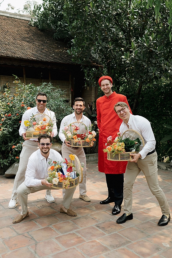 Wedding party portrait of groomsmen group photo holding wicker baskets of colorful flowers on a terracotta patio beside garden greenery and a rustic building
