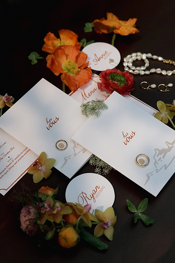 Wedding invitation suite flat lay with vow booklets, menu and place cards, rings, pearl jewelry, and orchid blooms on a dark tabletop