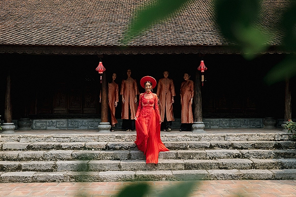 Bridal portrait of a bride in a red wedding dress with lace long sleeves and headpiece, descending stone steps at a temple entrance with red lanterns