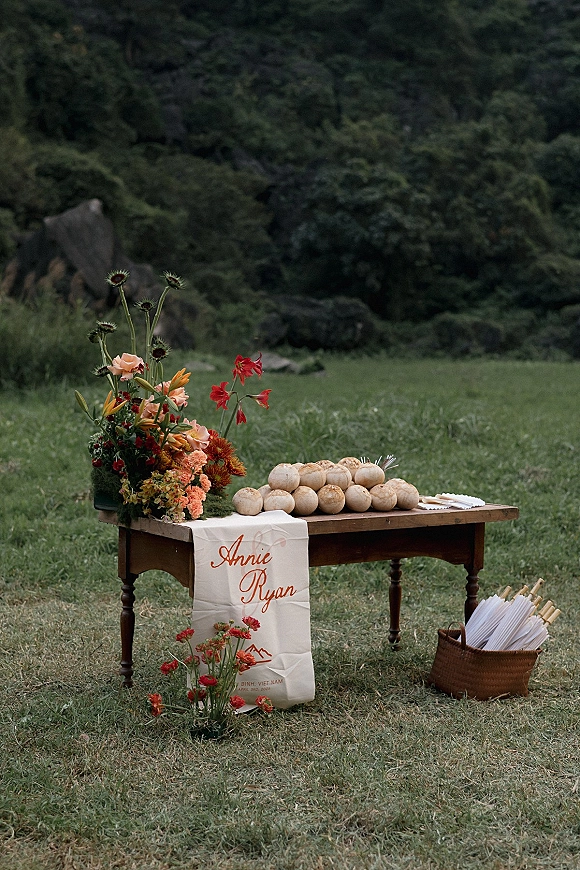 Wedding guest book table with calligraphy table runner, coconuts, paper parasols, and wildflower arrangement on a lawn by trees and rocks
