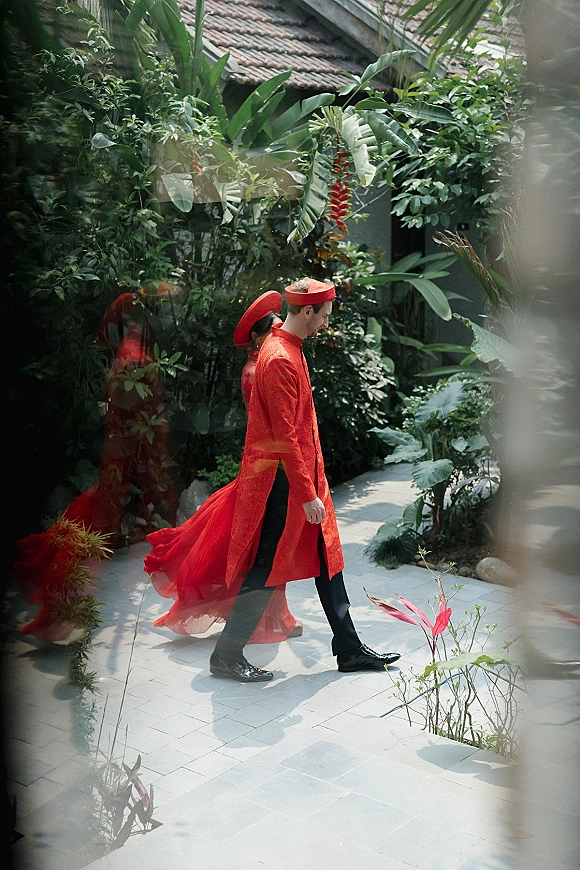 Wedding processional with bride and groom walking in traditional red wedding outfit along a tropical garden pathway with tiled walkway