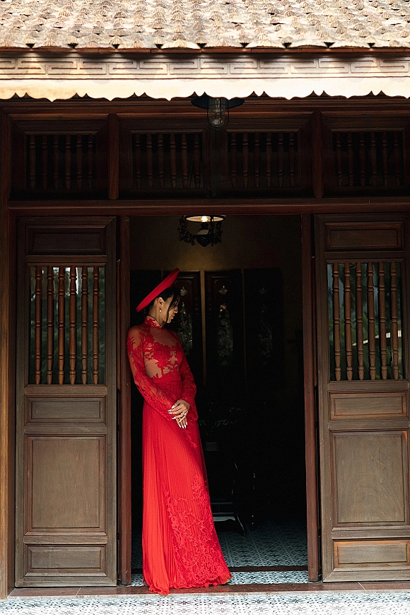Bridal portrait of a bride in a red wedding dress, standing in a wooden doorway of a traditional house with chandelier behind