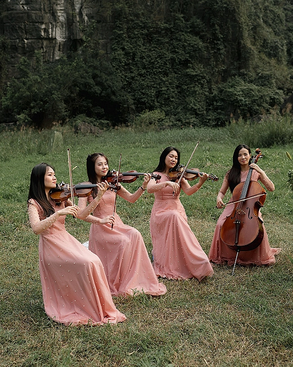 Wedding string quartet in blush dresses playing violins and cello with bows on a grass lawn beside greenery and a rock cliff