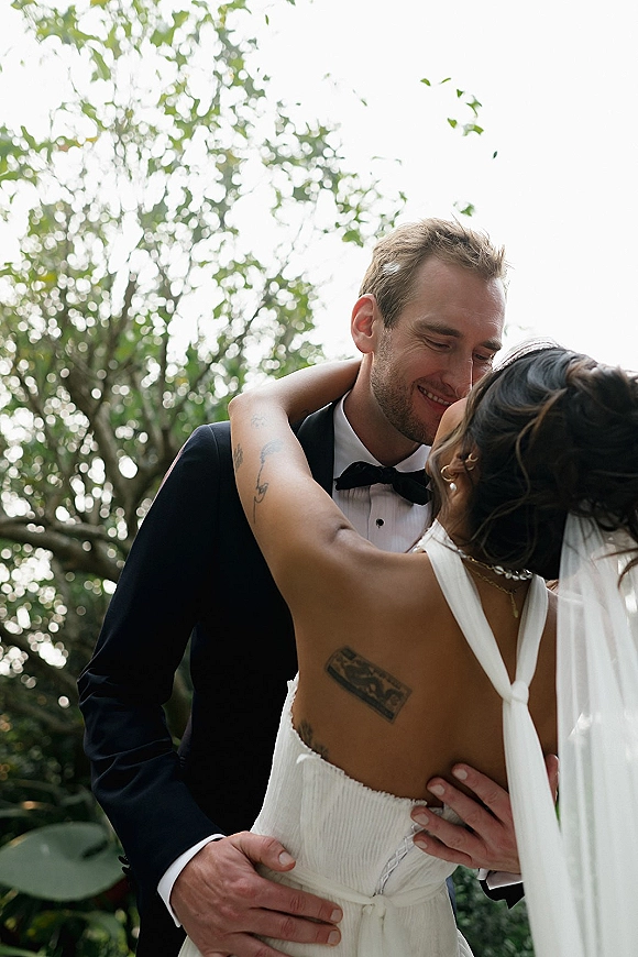 Wedding kiss portrait of bride and groom kissing, her veil and pearl earrings visible, his black tuxedo against leafy trees and sky