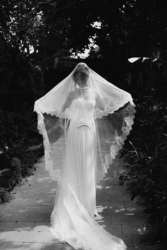 Bridal portrait of a bride holding her veil, lace edge framing her strapless corset gown and train on a dappled garden path