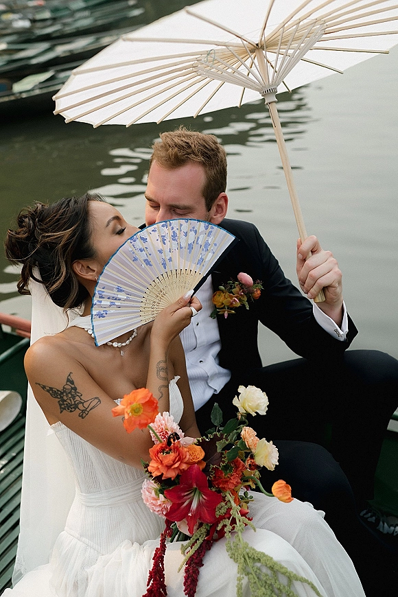 Wedding couple portrait of bride and groom in boat on calm water, bride in veil with orange-red bouquet and hand fan beside groom in tuxedo