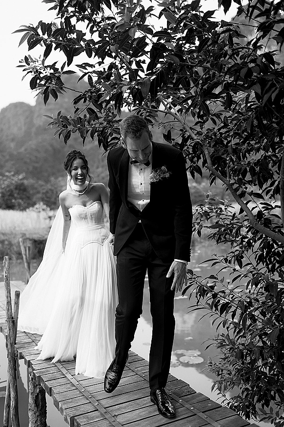 Couple portrait of bride and groom walking on a wooden dock by a lake, bride in strapless dress with veil, groom in tuxedo amid mountains