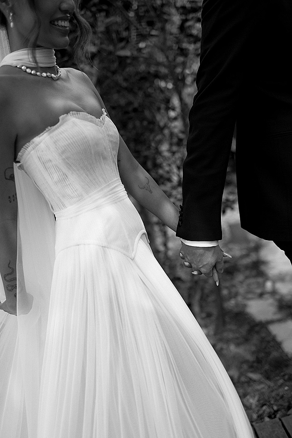 Couple holding hands with wedding rings close up, bride in strapless gown and veil beside groom in black suit on garden path