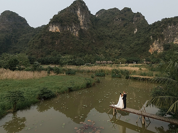Couple portrait of bride and groom on dock, her long veil and train flowing above a lily pad pond with mountain cliffs behind