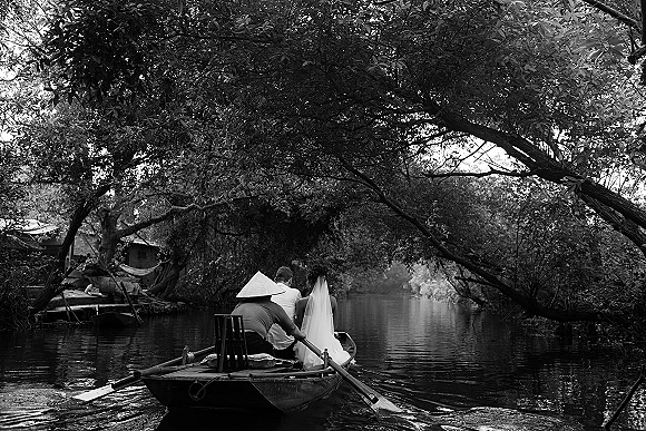 Couple portrait of bride and groom on boat, her long veil trailing as they row a river under overhanging trees and reflections