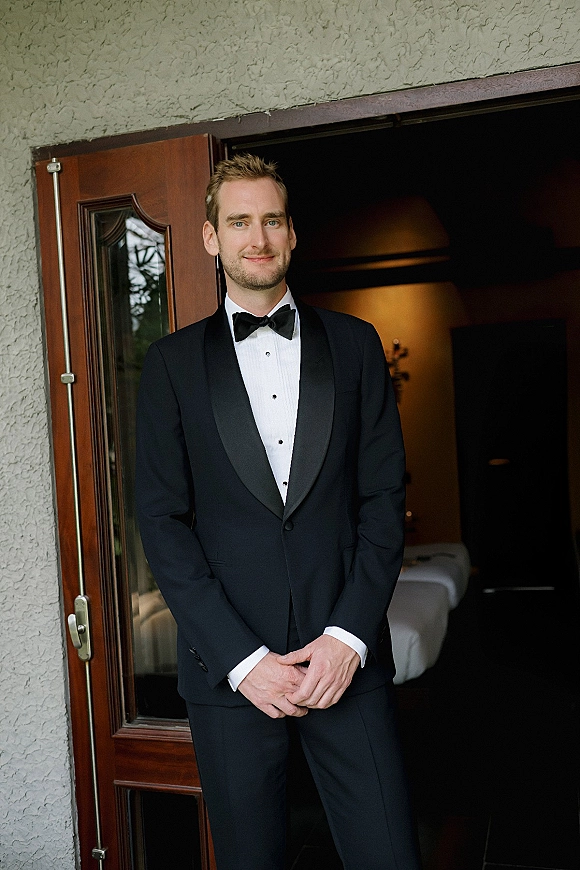 Groom portrait in a black tuxedo with bow tie and white dress shirt, hands clasped by a wood door under warm lamp light