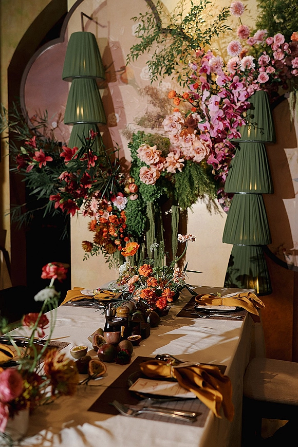 Reception tablescape with wedding table decor, lush orchid and rose centerpiece under hanging florals and pleated green pendant lamps by an arched niche