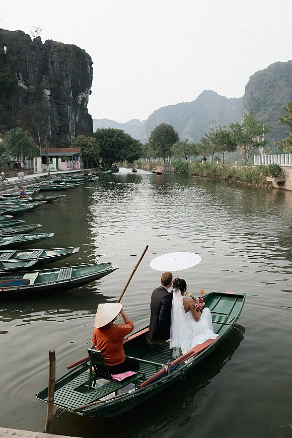 Couple portrait of bride and groom on boat with a white parasol and bouquet, drifting on a river beneath limestone cliffs and mountains