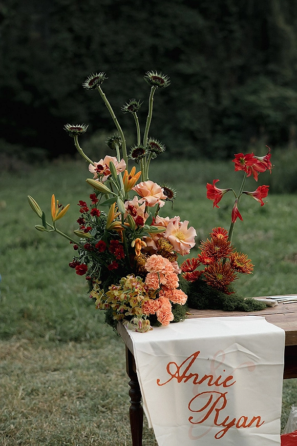 Wedding welcome sign on a wooden table with a fabric wedding sign, colorful florals and moss accents on a grassy lawn with trees