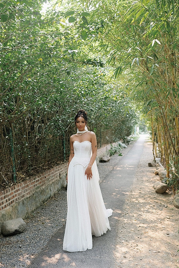 Bridal portrait in a strapless wedding dress, standing on a tree-lined bamboo path beside a brick wall with an updo hairstyle