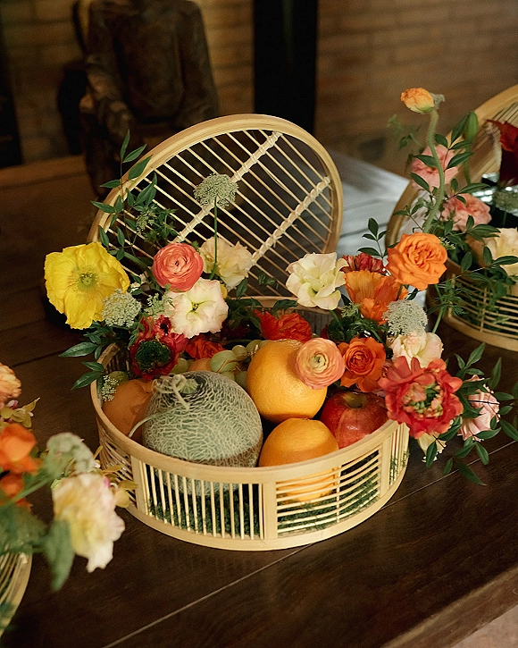 Wedding centerpiece with fruit wedding centerpiece styling, bright citrus and coral flowers in a wicker basket on a wood table by brick wall