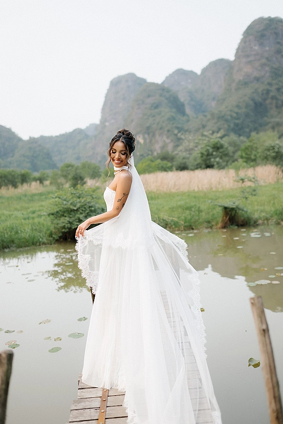 Bridal portrait of a bride on dock in a strapless wedding dress, holding a long lace-trim veil by a pond with lily pads and mountains
