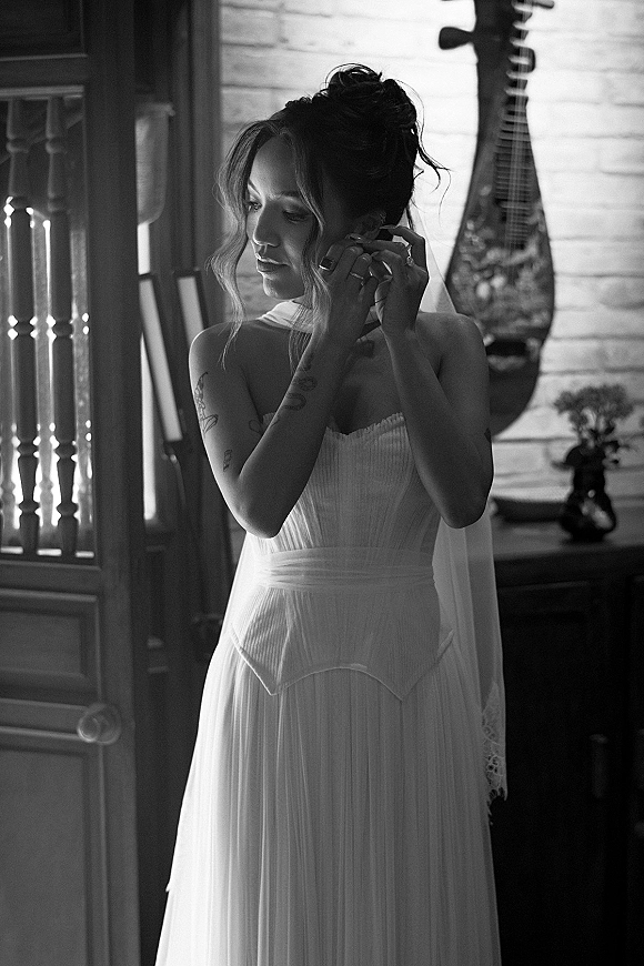 Bride getting ready in window light, adjusting earrings with veil over shoulders and wedding dress beside a brick wall in black and white