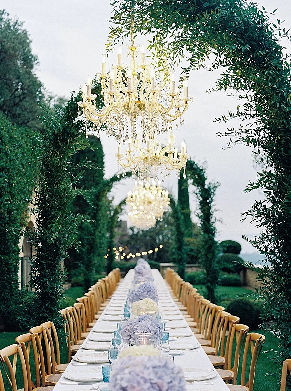 Outdoor reception tablescape with a garden banquet table set in white linens, hydrangea centerpieces, votive candles, and crystal chandeliers under string lights