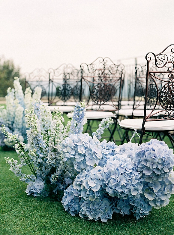 Ceremony aisle decor with blue hydrangea aisle flowers and delphinium arrangements lining wrought iron chairs on a grassy lawn under open sky