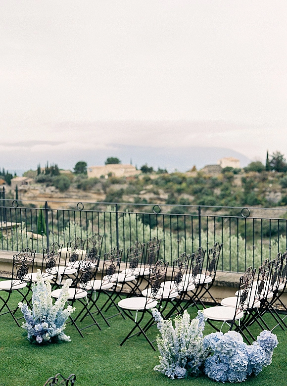 Ceremony seating with outdoor ceremony chairs in wrought iron rows, blue hydrangea accents lining the aisle on a terrace lawn with hills beyond
