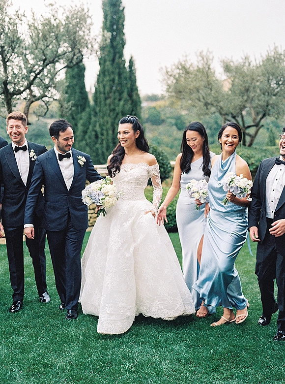 Wedding party portrait of bride and groom with bridesmaids and groomsmen walking on a garden lawn, bride in off-shoulder lace gown holding bouquet