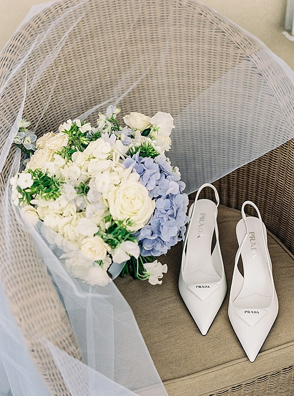 Bridal shoes, white slingback wedding heels on a wicker chair with tulle veil, white roses, and blue hydrangea bouquet accents