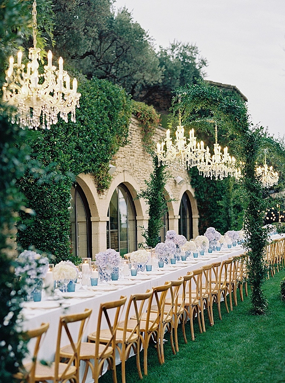 Outdoor reception tablescape with a long banquet table wedding setup, hydrangea centerpieces and votive candles beneath crystal chandeliers by a stone facade