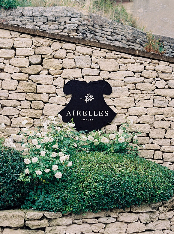 Wedding venue sign with white lettering and floral emblem on a black board, framed by white roses and greenery against a stone wall backdrop