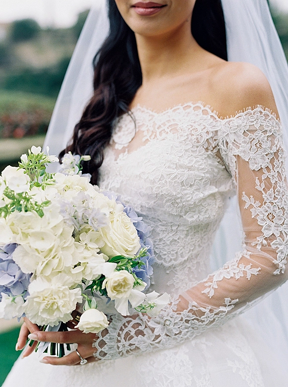 Bridal portrait of a bride holding bouquet of white roses and blue hydrangea, wearing an off-the-shoulder lace gown with cathedral veil in a blurred garden