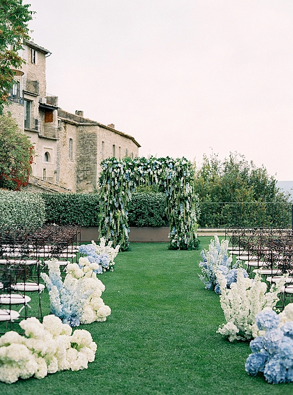 Ceremony setup for an outdoor wedding ceremony with a floral arch and blue hydrangea aisle flowers on a garden lawn by a stone villa