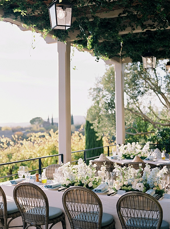 Reception tablescape with white linens and floral centerpieces, greenery garland, blue napkins and glassware under a vine-covered pergola on a terrace