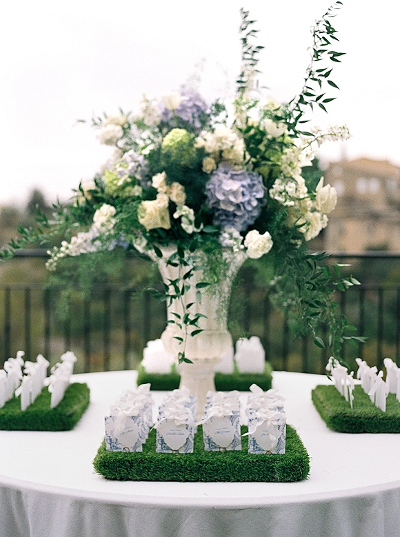 Wedding escort cards arranged in ribbon-tied favor boxes on faux grass trays beneath a tall hydrangea centerpiece on an outdoor terrace table