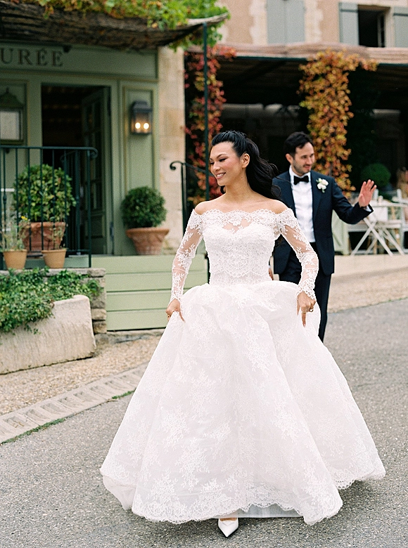 Bride portrait in an off the shoulder lace wedding dress twirling beside groom in tuxedo on a street with green storefront and ivy